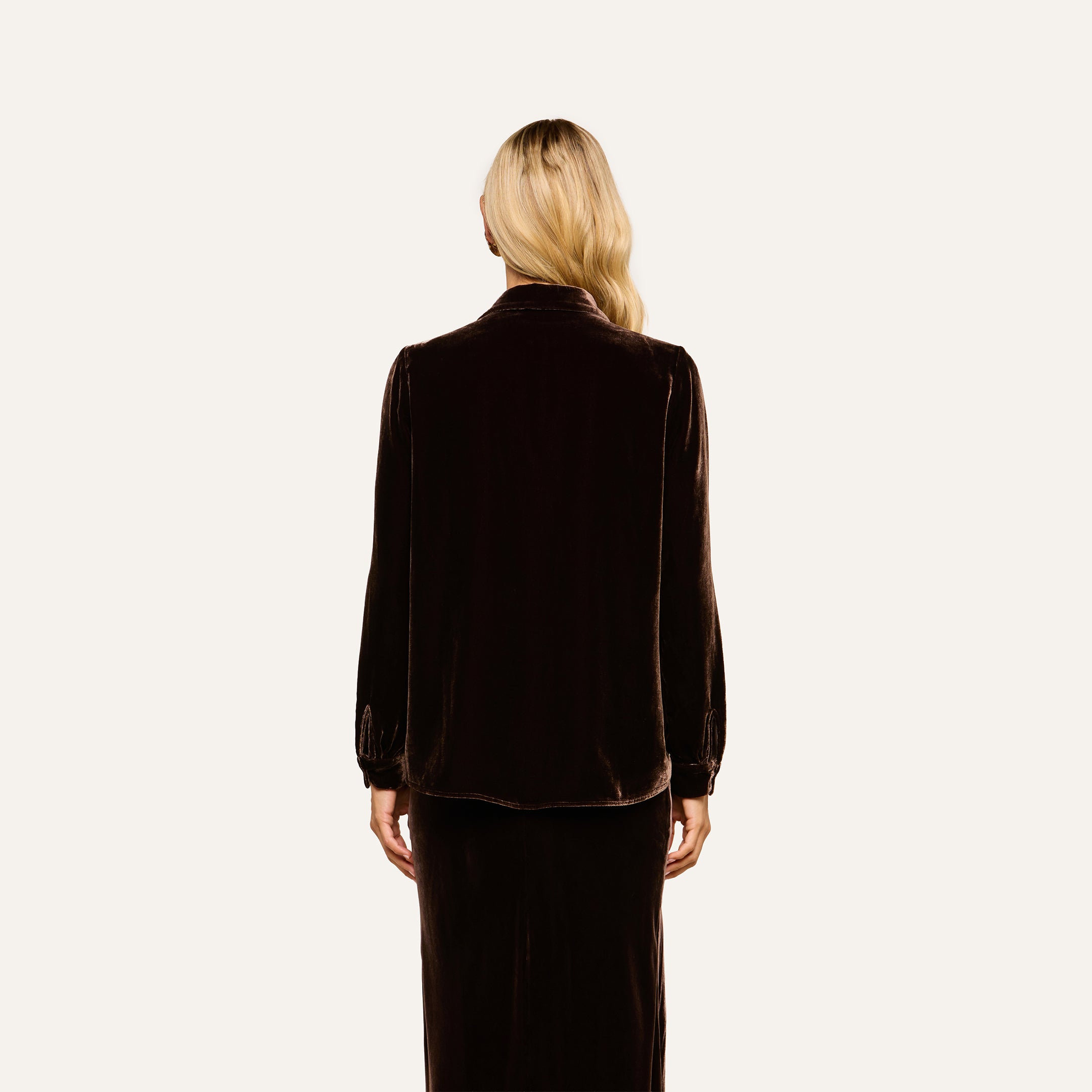 A woman with long blonde hair stands facing away, wearing the Argo Velvet Blouse in dark java brown with long sleeves and a matching skirt against a plain light background.