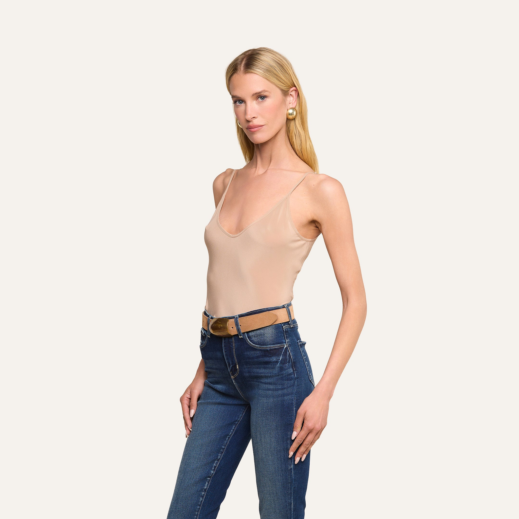 Une femme aux longs cheveux blonds porte le caraco Lexi Silk de couleur beige, assorti dun jean bleu et de boucles doreilles dorées ; elle pose, une main sur la hanche, devant un fond beige uni.