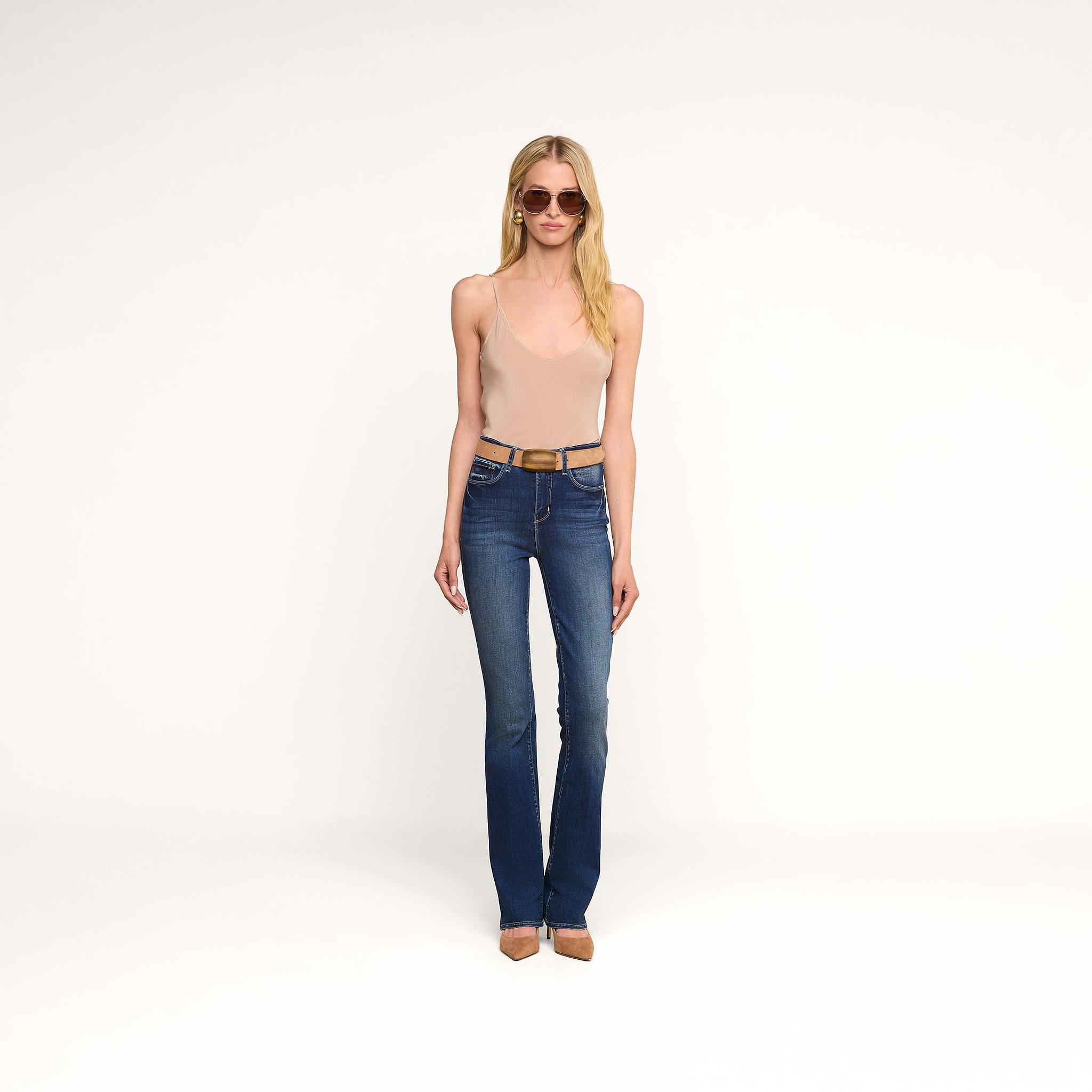 Une femme portant des lunettes de soleil, le caraco « Lexi Silk » de couleur beige et un jean bootcut bleu, avec une ceinture et des chaussures marron, se tenant debout devant un fond clair uni.