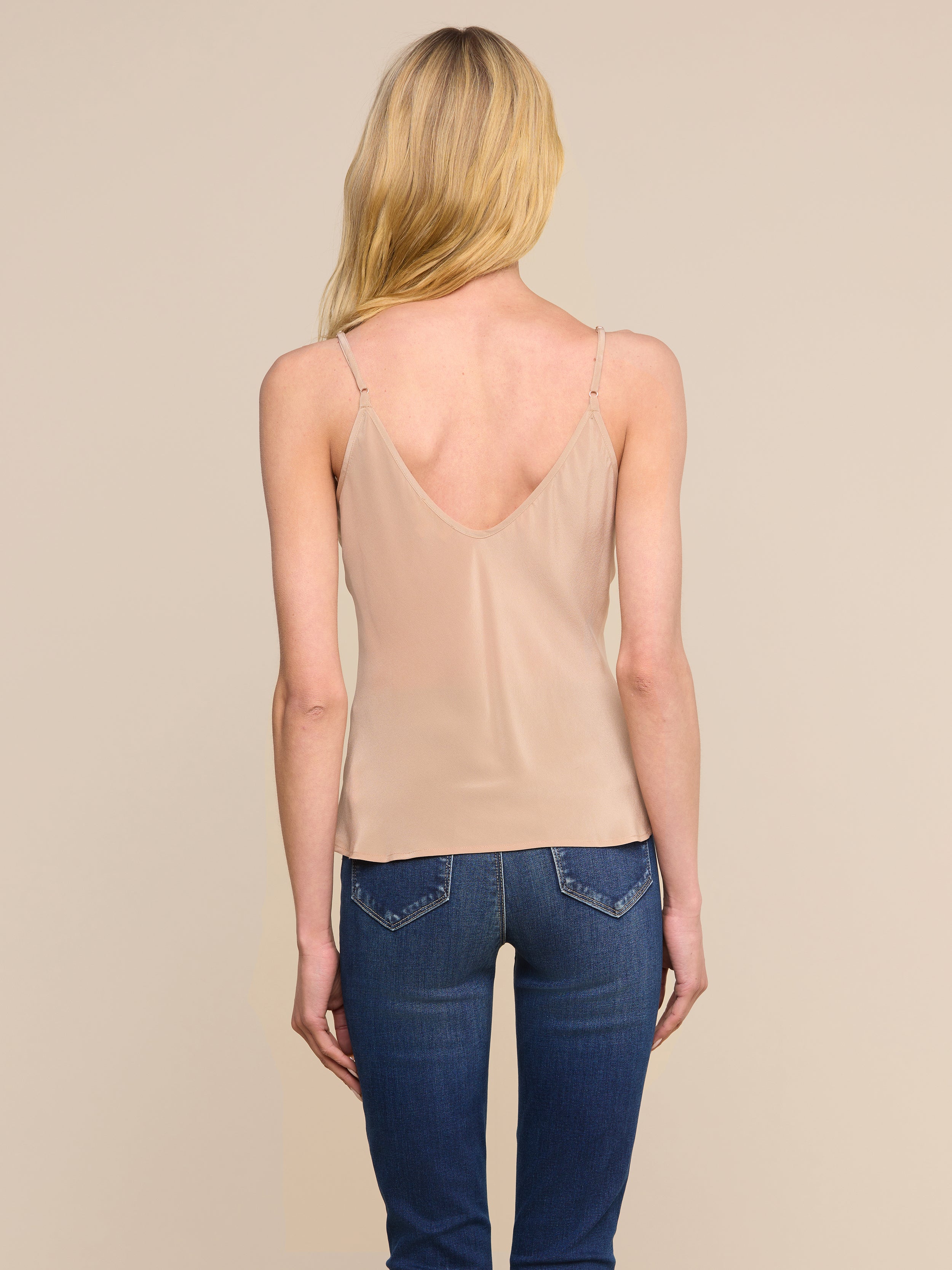 On voit de dos une femme aux longs cheveux blonds, vêtue dun caraco Lexi Silk beige et dun jean bleu, debout devant un fond beige uni.