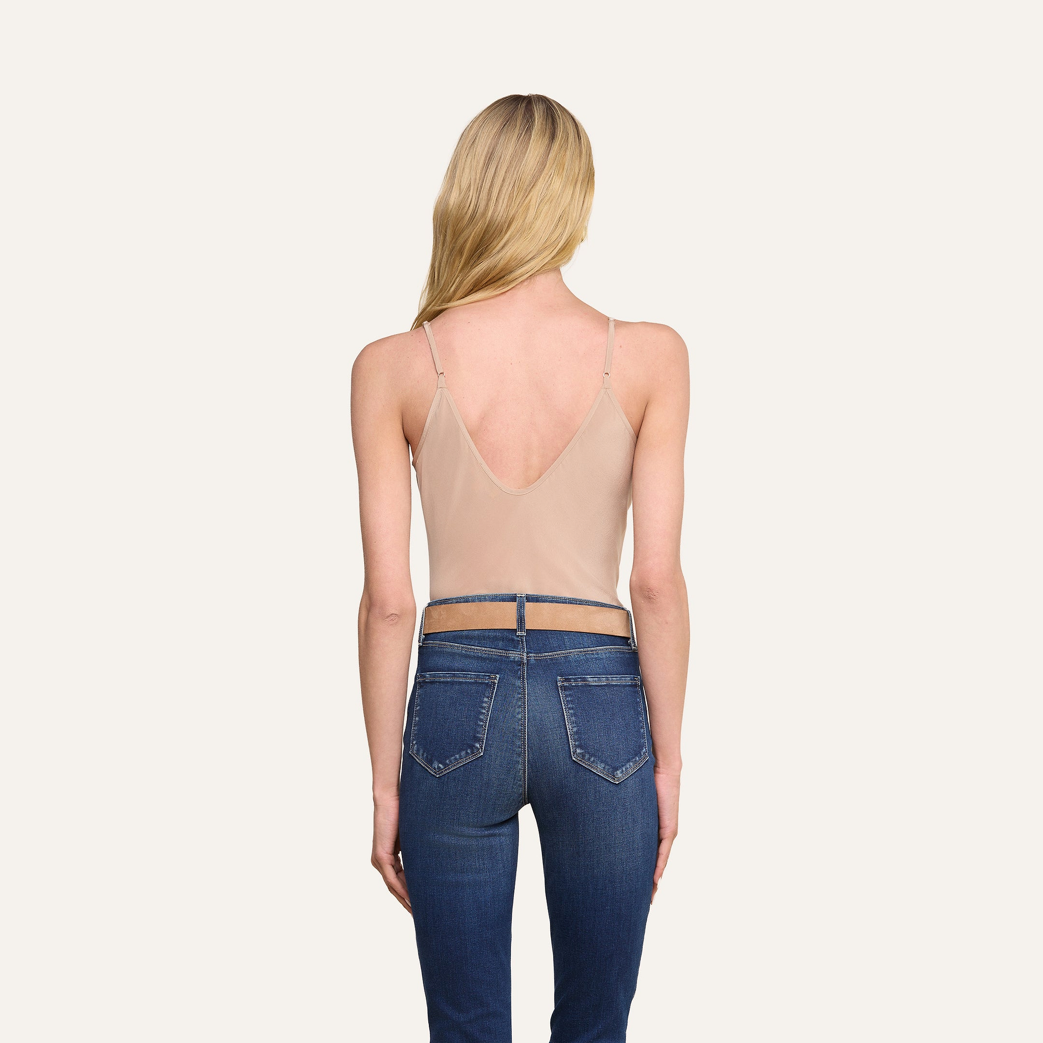 Une femme aux longs cheveux blonds, vue de dos, porte le caraco Lexi Silk — un haut couleur chair à coupe en biais doté de bretelles réglables — associé à un jean bleu taille haute, sur un fond beige uni.