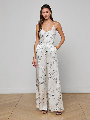 A woman stands indoors against a plain wall, wearing the Lexi Satin Camisole—a white sleeveless top with black abstract patterns and adjustable straps. She has long brown hair and her hands in her pockets.