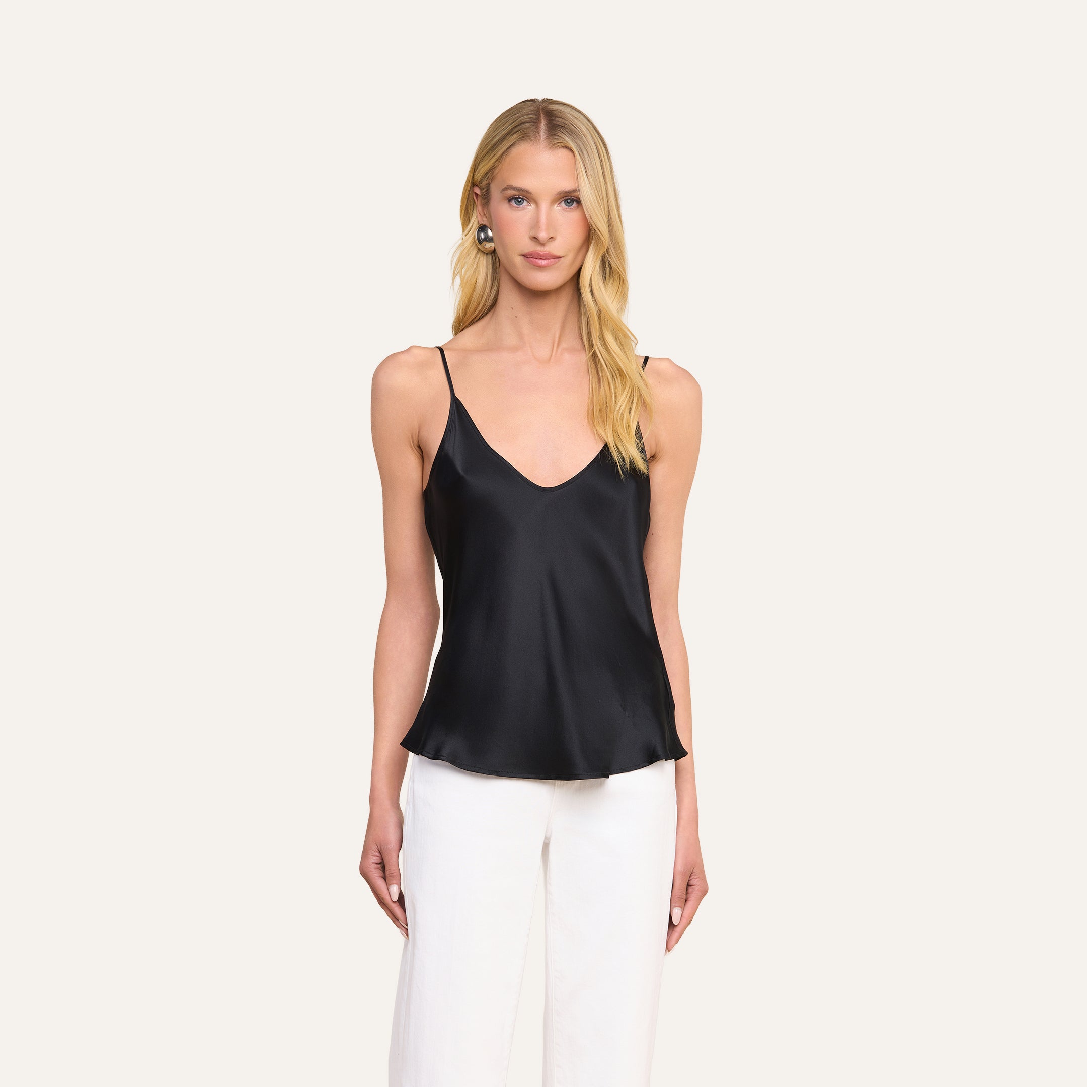 A woman stands against a plain light background, facing forward with her arms at her sides, wearing the Lexi Silk Camisole in black with adjustable spaghetti straps and white pants.