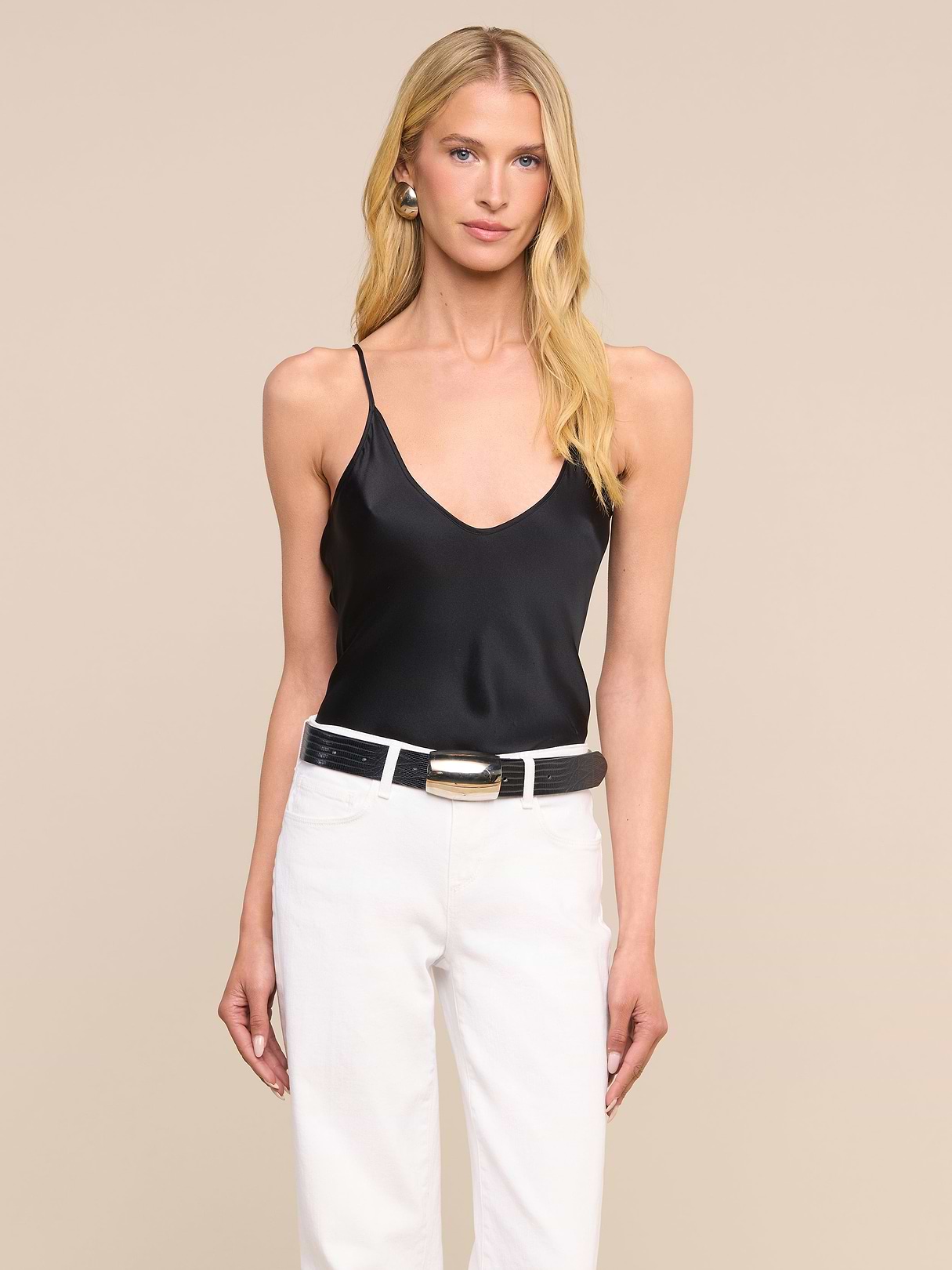 Une femme aux longs cheveux blonds porte le caraco en soie Lexi, doté de bretelles fines réglables, associé à un pantalon blanc, une ceinture noire à boucle dorée et des boucles doreilles créoles, debout devant un fond beige uni.