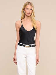 A woman with long blonde hair stands against a plain light background, wearing the Lexi Silk Camisole with adjustable spaghetti straps, paired with white pants, a black belt, and earrings.