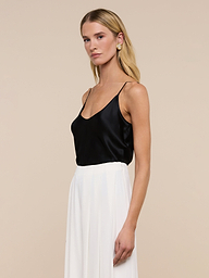 A woman with long blonde hair wears the Lexi Silk Camisole—a black silk top with adjustable spaghetti straps—paired with white high-waisted pants, a black belt, and statement earrings. She stands against a plain light background, looking slightly to her right with a neutral expression.