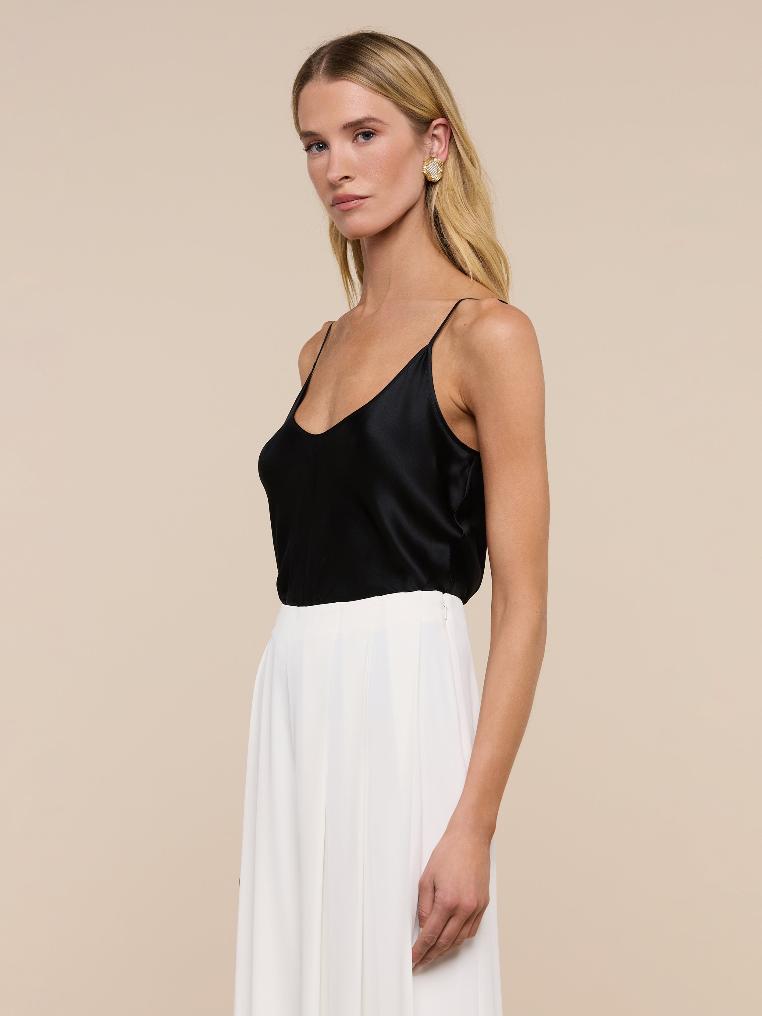 A woman with long blonde hair wears the Lexi Silk Camisole—a black silk top with adjustable spaghetti straps—paired with white high-waisted pants, a black belt, and statement earrings. She stands against a plain light background, looking slightly to her right with a neutral expression.