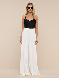 A woman wearing sunglasses, the Lexi Silk Camisole with adjustable spaghetti straps, white wide-leg pants, a black belt, and black heels stands against a plain white background.