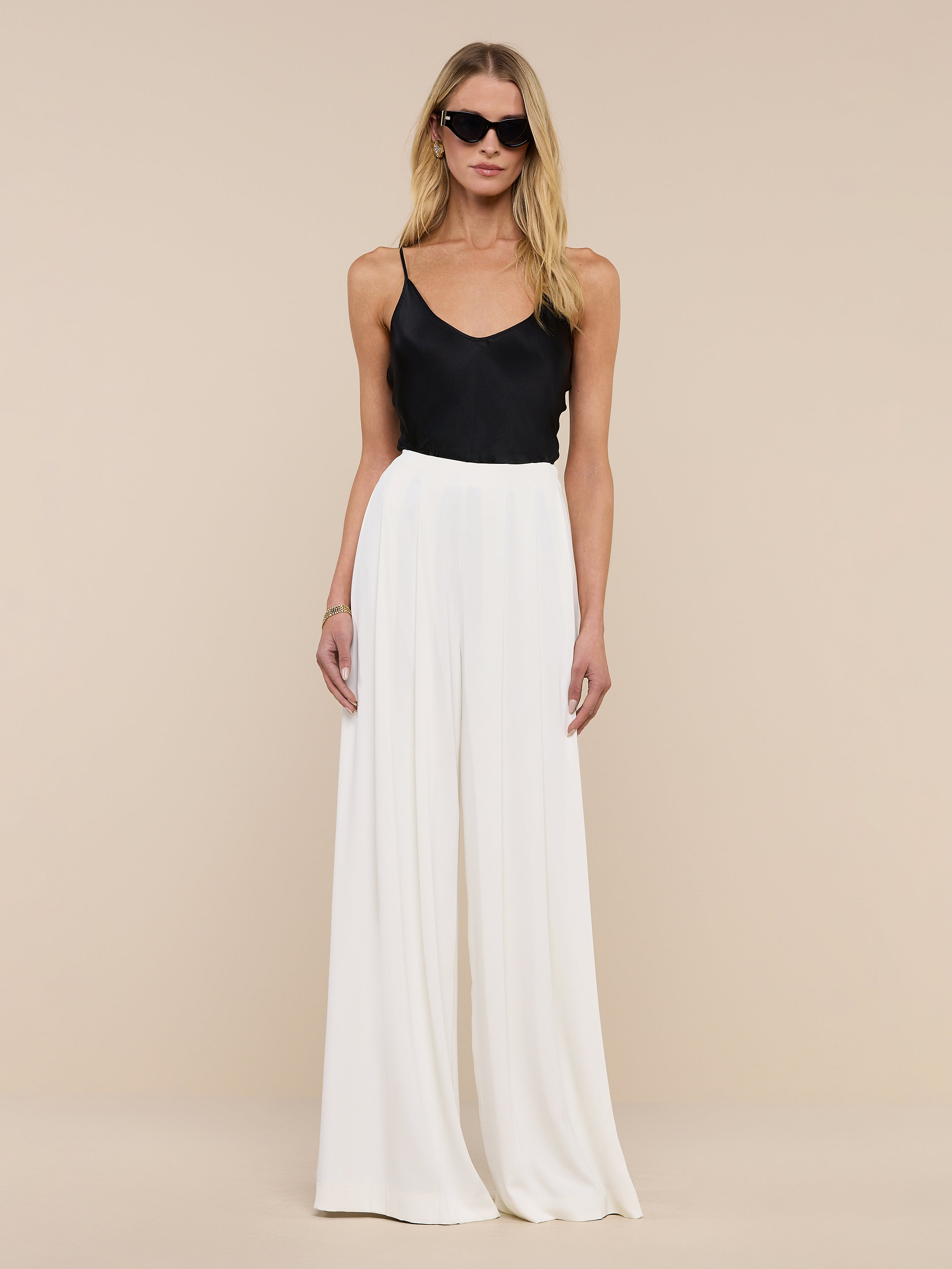 A woman wearing sunglasses, the Lexi Silk Camisole with adjustable spaghetti straps, white wide-leg pants, a black belt, and black heels stands against a plain white background.