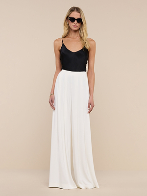 Wearing black sunglasses and the Lexi Silk Camisole with adjustable spaghetti straps, a woman pairs it with high-waisted wide-leg white pants, standing against a plain beige background, facing forward with arms at her sides.