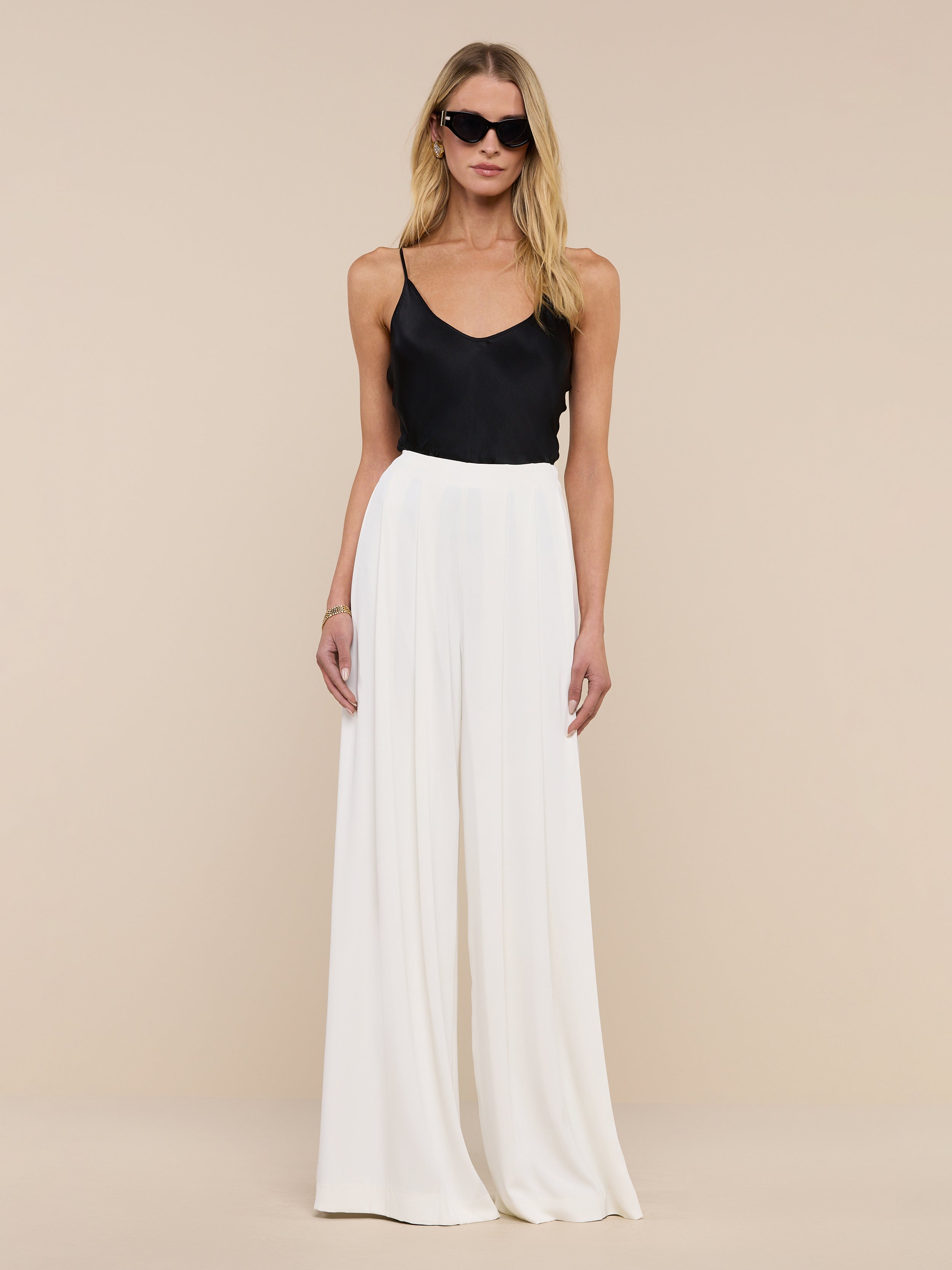 A woman wearing sunglasses, the Lexi Silk Camisole with adjustable spaghetti straps, white wide-leg pants, a black belt, and black heels stands against a plain white background.