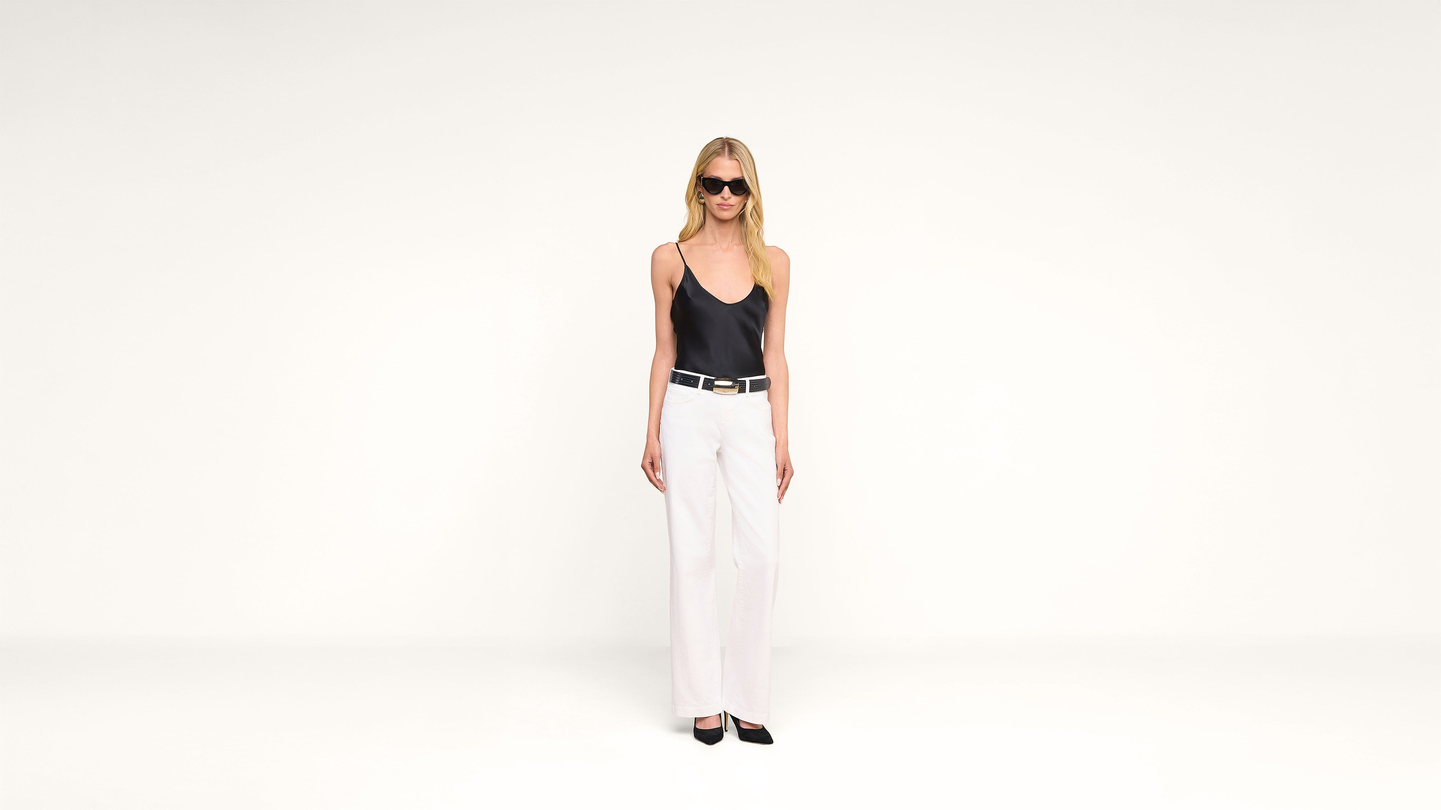 A woman wearing sunglasses, a black tank top, white pants, and black shoes stands against a plain white background.