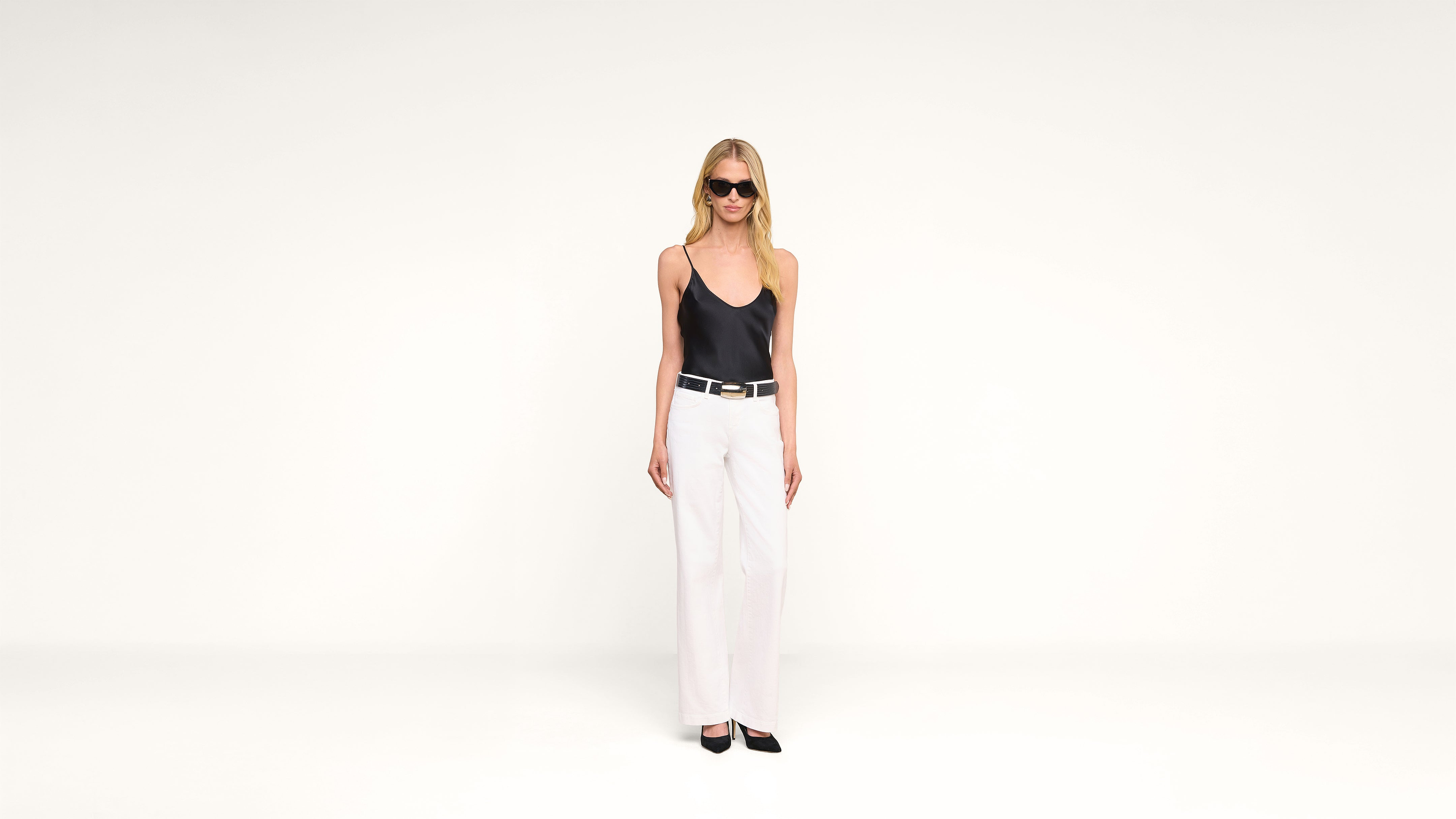A woman wearing sunglasses, a black tank top, white pants, and black shoes stands against a plain white background.