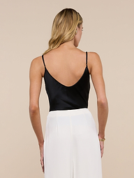 A woman with long blonde hair is seen from behind, standing against a plain light background, wearing the Lexi Silk Camisole in black with adjustable spaghetti straps and white pants.