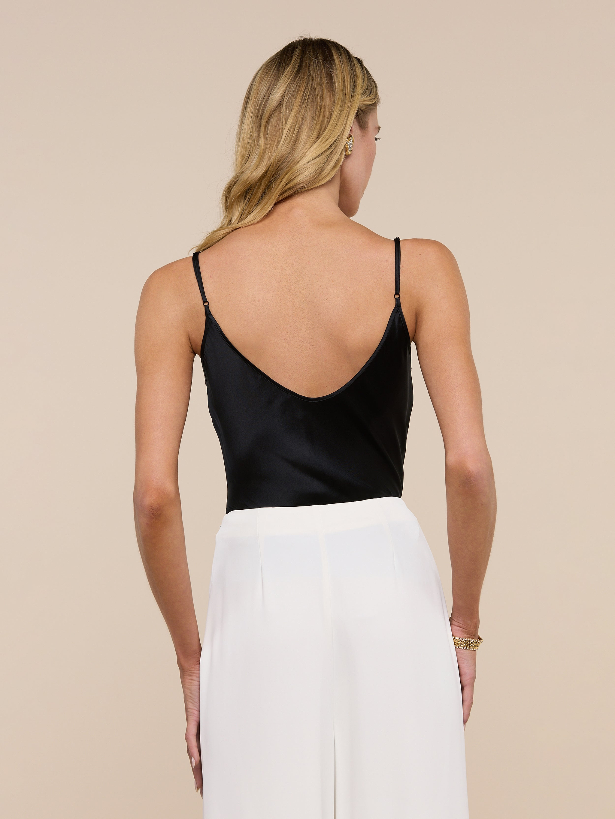 A woman with long blonde hair is seen from behind, standing against a plain light background, wearing the Lexi Silk Camisole in black with adjustable spaghetti straps and white pants.