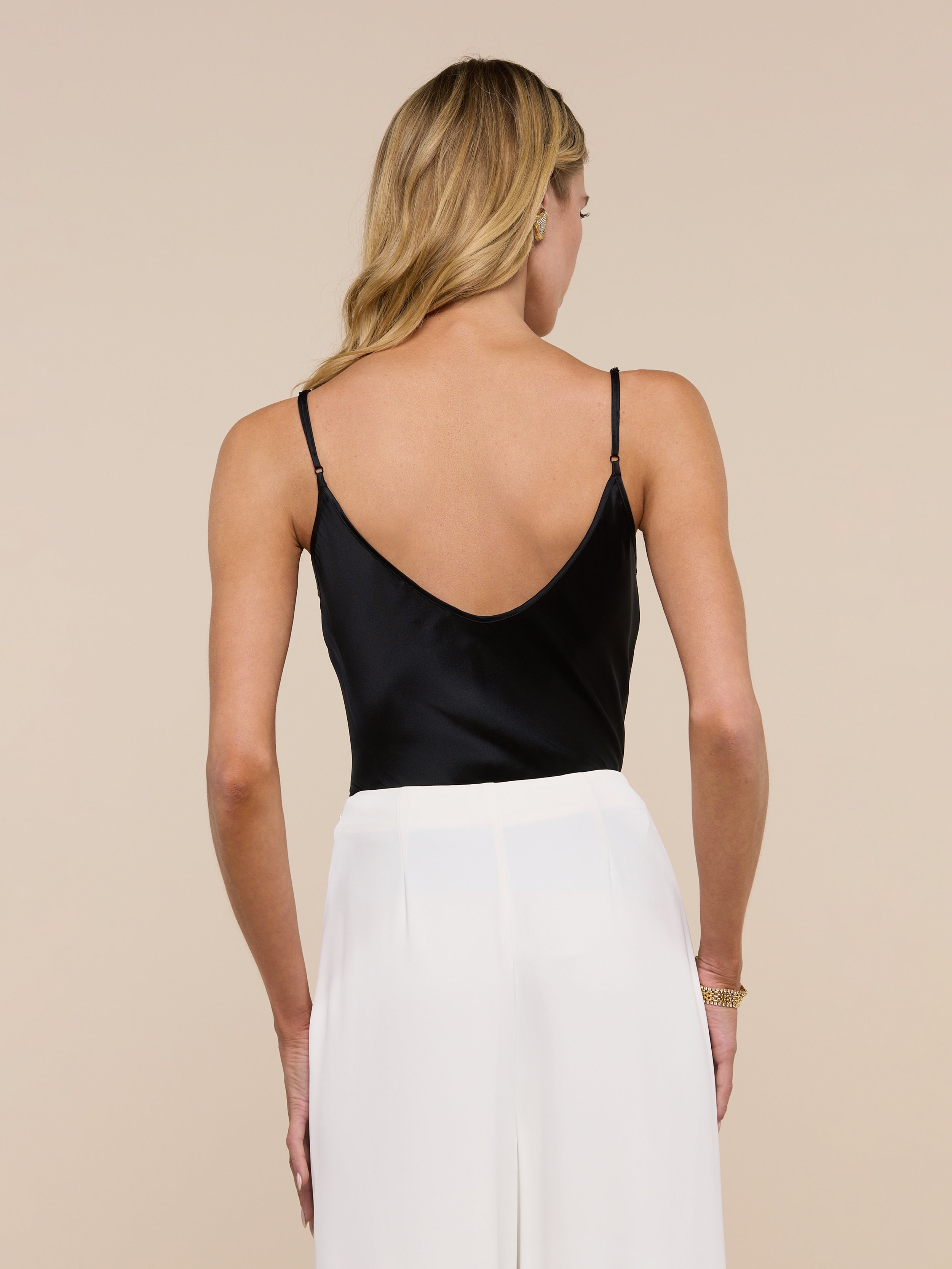 A woman with long blonde hair is seen from behind, standing against a plain light background, wearing the Lexi Silk Camisole in black with adjustable spaghetti straps and white pants.