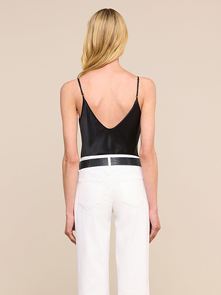 Une femme aux longs cheveux blonds, vue de dos, porte le caraco Lexi Silk noir à bretelles fines réglables, associé à un pantalon blanc taille haute et à une ceinture noire. Larrière-plan est dun beige uni.