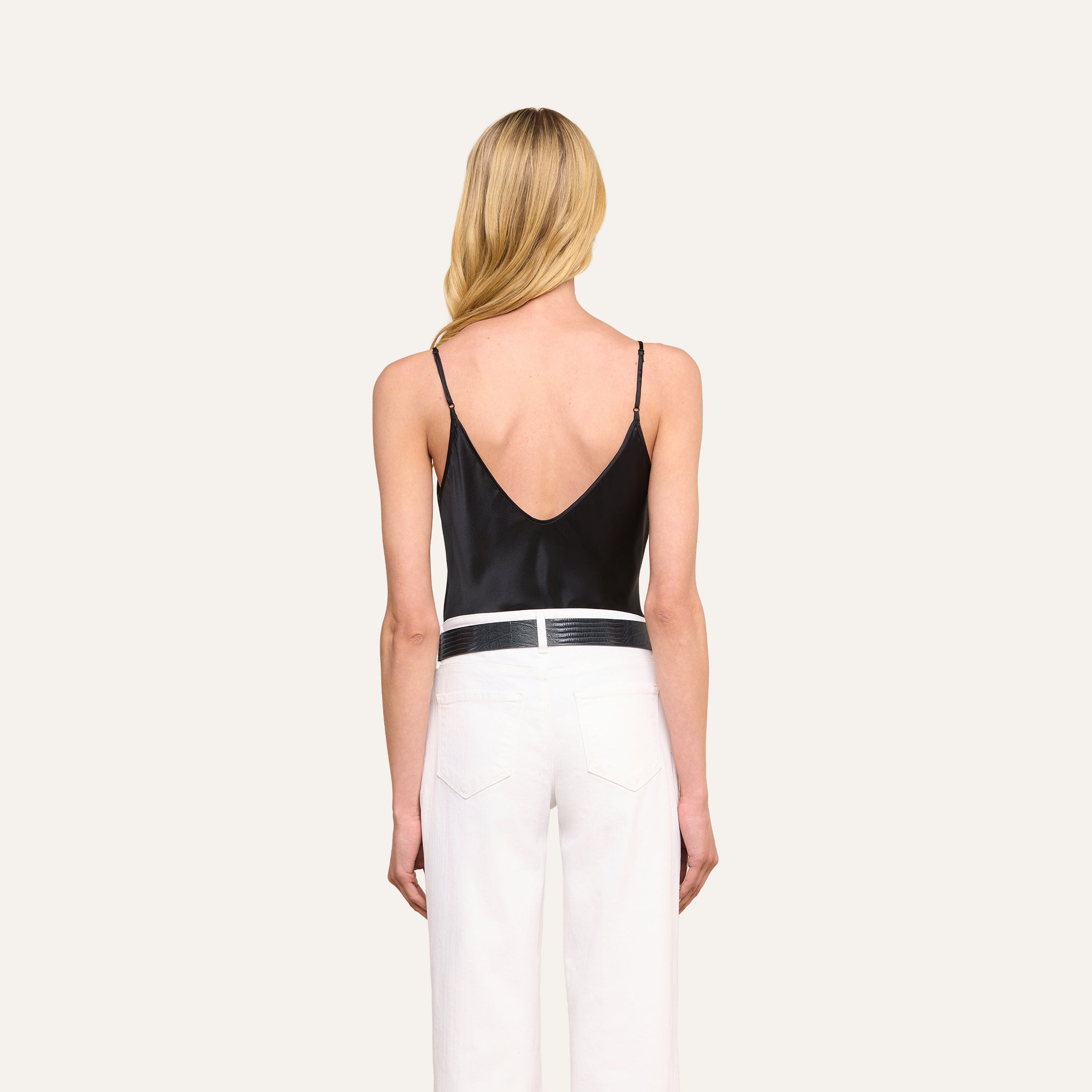 A woman with long blonde hair, dressed in the Lexi Silk Camisole with adjustable spaghetti straps and white pants, stands with her back to the camera against a plain light background.