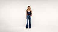 A woman stands against a plain white background wearing a black sleeveless top, blue flared jeans with a tan belt, and beige shoes.