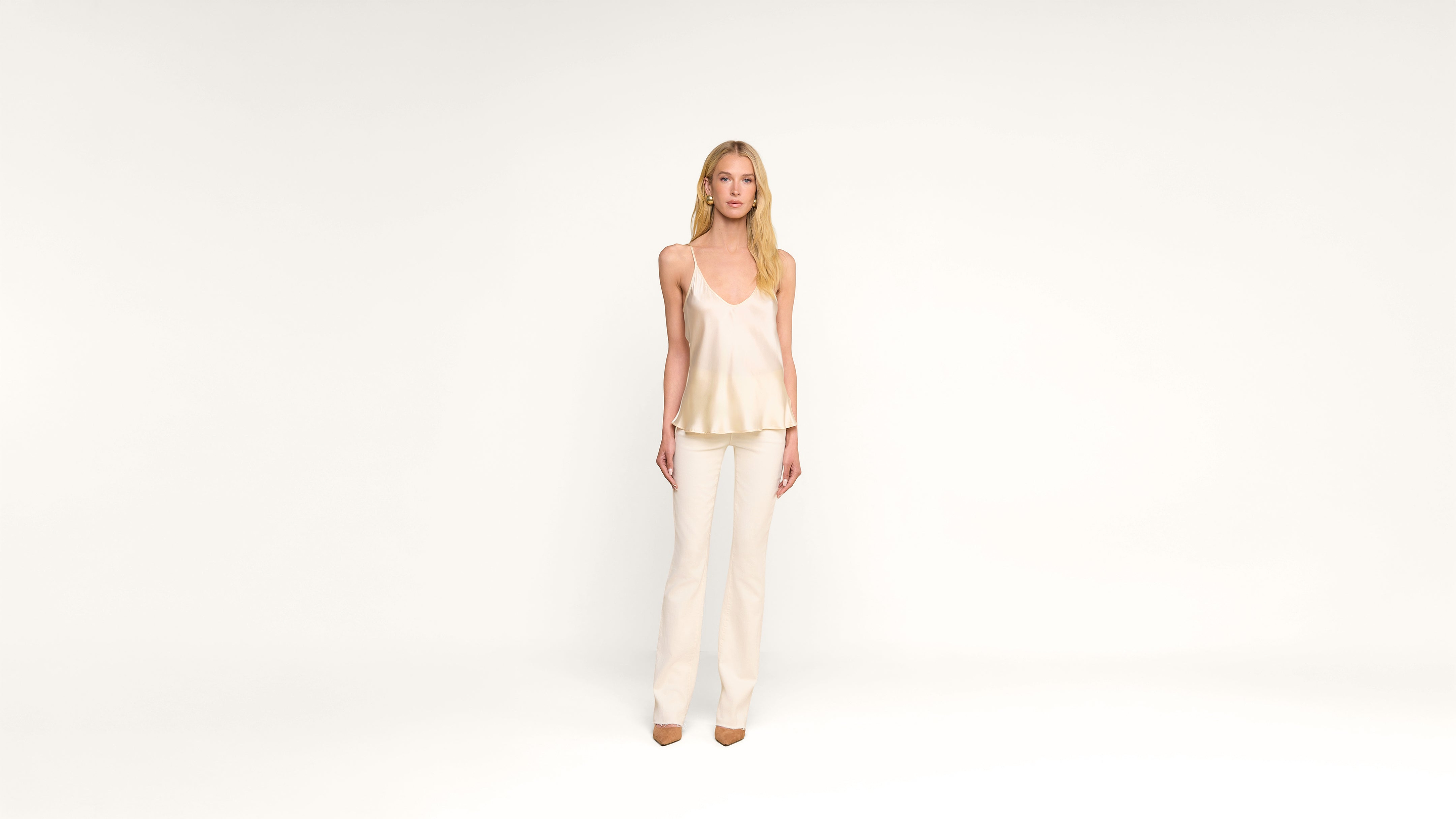 A woman with long blonde hair stands against a plain white background, wearing a cream-colored sleeveless top, cream pants, and beige shoes. She faces forward with a neutral expression.
