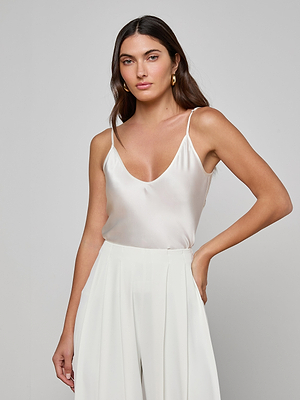 A woman with long brown hair wears the Lexi Silk Camisole, featuring adjustable straps, and high-waisted white trousers, standing against a plain light gray background with a neutral expression.