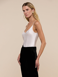 A woman with long blonde hair wears the Lexi Silk Camisole, a sleeveless white charmeuse top with adjustable straps, paired with black pants. She stands against a plain light background, looking slightly to the side.