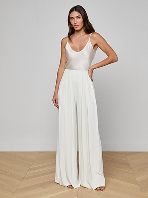 A woman with long, loose brown hair poses indoors against a plain white wall and light wood floor, wearing the Lexi Silk Camisole with adjustable straps and high-waisted, wide-leg white trousers.