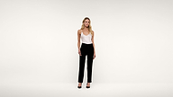 A woman stands against a plain white background wearing a white sleeveless top, black pants, and black heels, with her arms at her sides.
