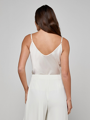 A woman with long brown hair is seen from behind, wearing the Lexi Silk Camisole with adjustable straps and white high-waisted pants, set against a plain light gray background.