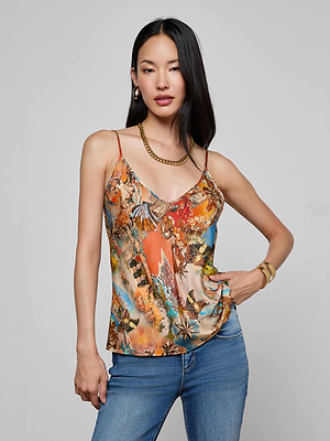 A woman with long straight black hair poses against a plain background, wearing the Lexi Satin Camisole—a colorful bias-cut satin top with adjustable straps—paired with blue jeans and gold jewelry. She stands relaxed, one hand on her hip.