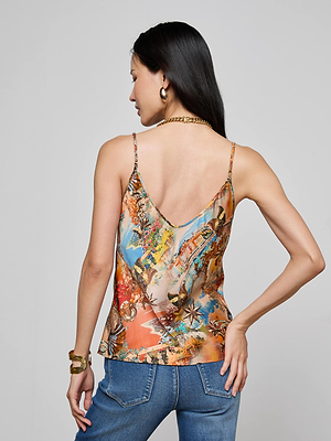 A woman with long dark hair, wearing gold jewelry and the Lexi Satin Camisole in a travel postcard print with adjustable straps, stands with her back to the camera and left hand on her hip against a plain light background.