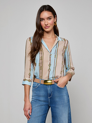 A woman with long brown hair stands against a plain background, wearing the Camille Blouse in beige and light blue stripes, paired with gold hoop earrings and blue jeans. She has one hand in her pocket and is smiling slightly.