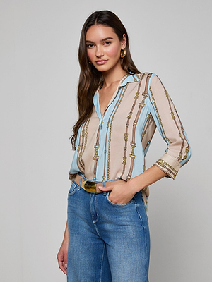 A woman with long brown hair wears the Camille Blouse, featuring three-quarter sleeves and a striped belt print, paired with blue jeans and gold hoop earrings. She stands smiling slightly with one hand in her pocket against a plain background.