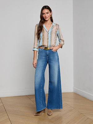 A woman with long brown hair stands indoors on a wooden floor, smiling slightly. She wears the Camille Blouse, a three-quarter sleeve striped belt print shirt, with wide-leg blue jeans, a brown belt, heeled boots, and gold earrings.