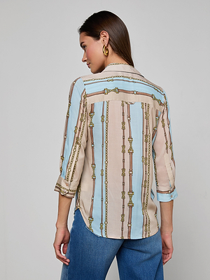 A woman with long brown hair faces away, wearing the Camille Blouse—a viscose twill top with three-quarter rolled sleeves and a beige and blue stripe-and-chain print. She pairs it with blue jeans and gold hoop earrings.