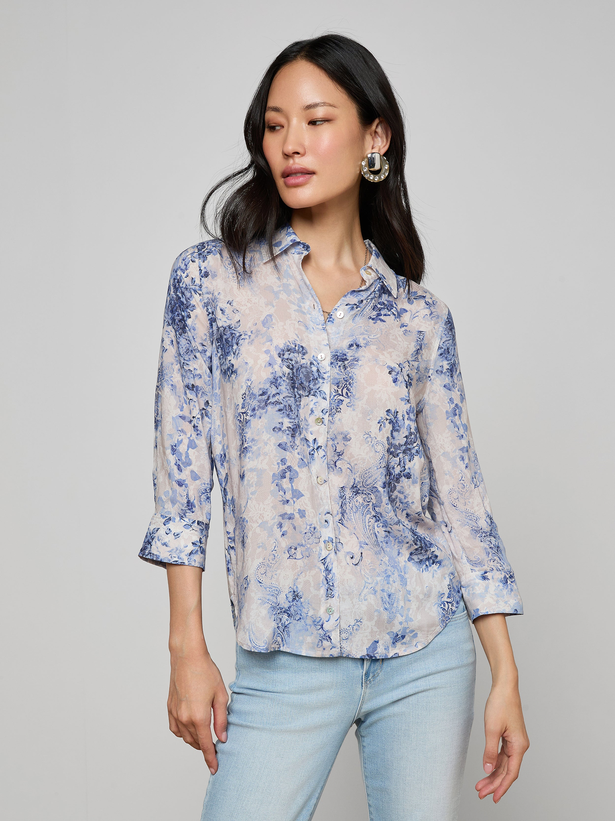 A woman stands against a plain background, one hand by her side and the other slightly raised, wearing the Camille Blouse—a light, three-quarter sleeve top crafted from viscose twill with an ivory and blue lace print—paired with light blue jeans.