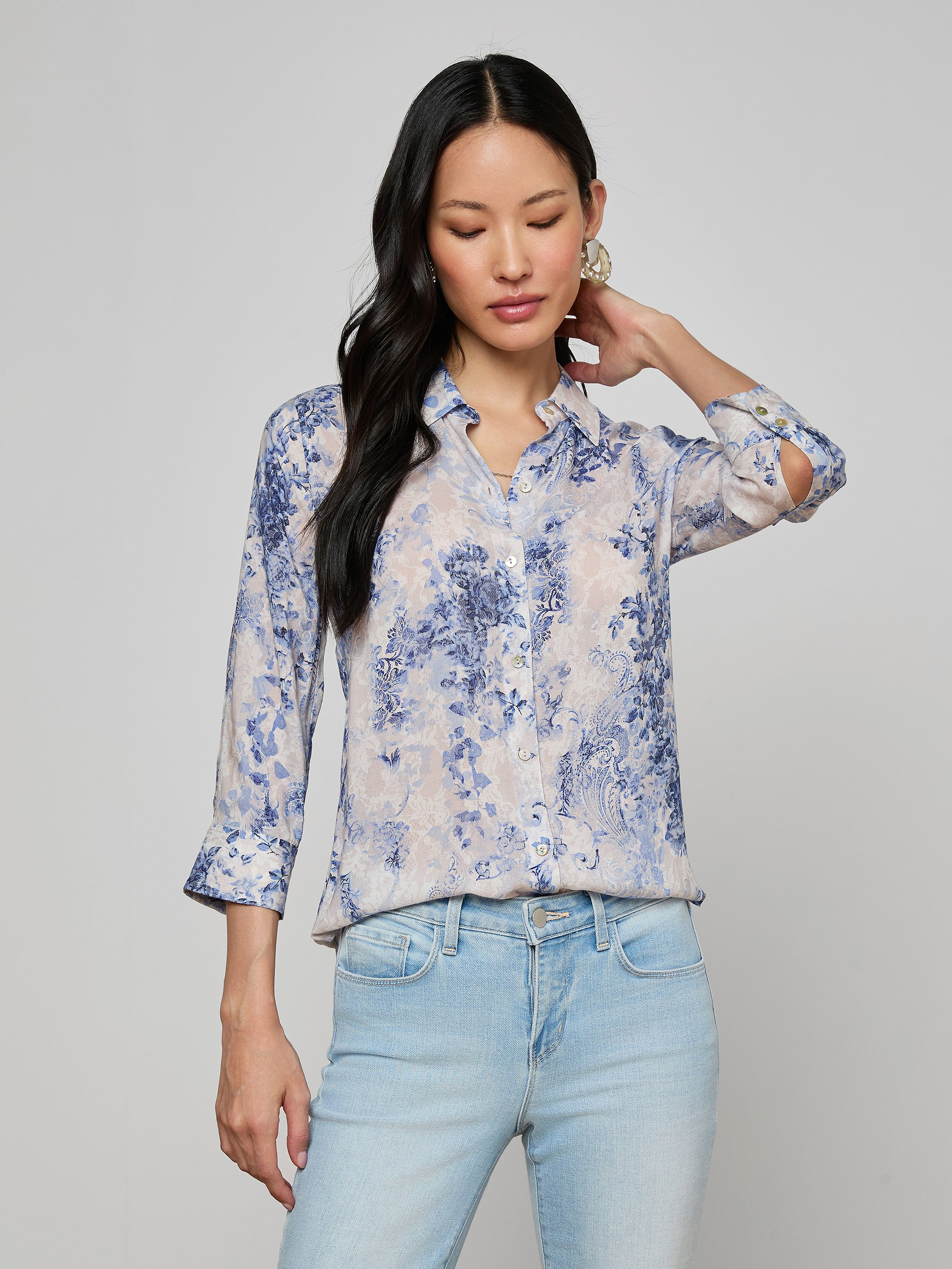 A woman stands against a plain background, looking down with one hand touching her hair, wearing the Camille Blouse—an ivory and blue lace print three-quarter sleeve shirt made from viscose twill—paired with light blue jeans.