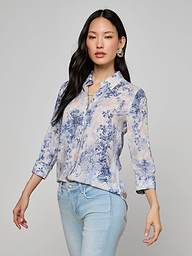A woman wearing the Camille Blouse—a light blue and white floral button-up shirt with three-quarter sleeves, crafted from viscose twill featuring an ivory and blue lace print—paired with light blue jeans, stands against a plain background.