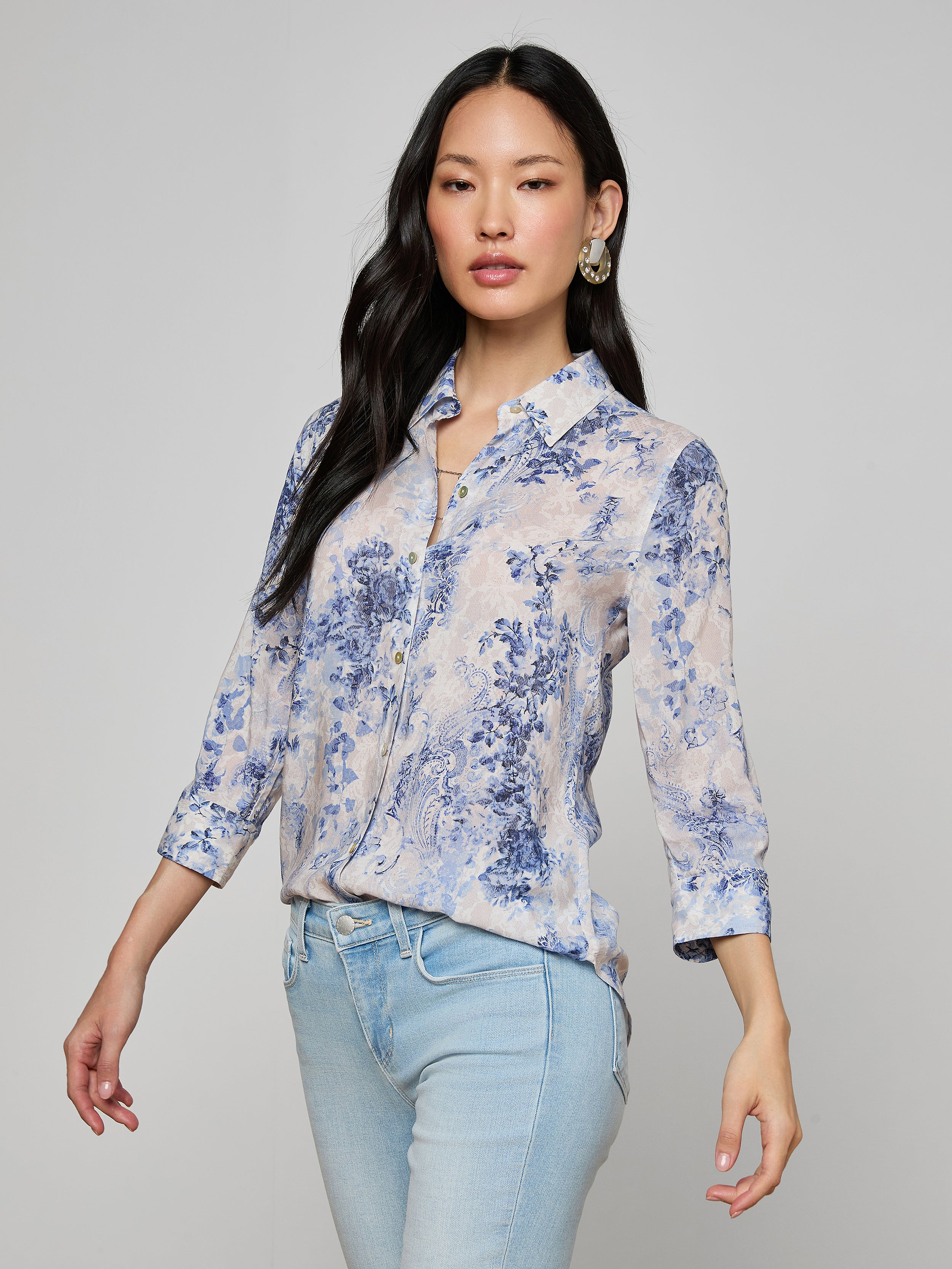 A woman wearing the Camille Blouse—a light blue and white floral button-up shirt with three-quarter sleeves, crafted from viscose twill featuring an ivory and blue lace print—paired with light blue jeans, stands against a plain background.
