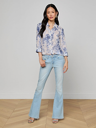 Woman standing against a plain background, wearing the Camille Blouse—an ivory and blue lace print three-quarter sleeve shirt crafted from viscose twill—paired with light-wash flared jeans and open-toe heels. She has long dark hair and a relaxed, confident pose.