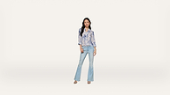 A woman stands against a plain white background, wearing a light blue floral blouse, light blue flared jeans, and sandals.