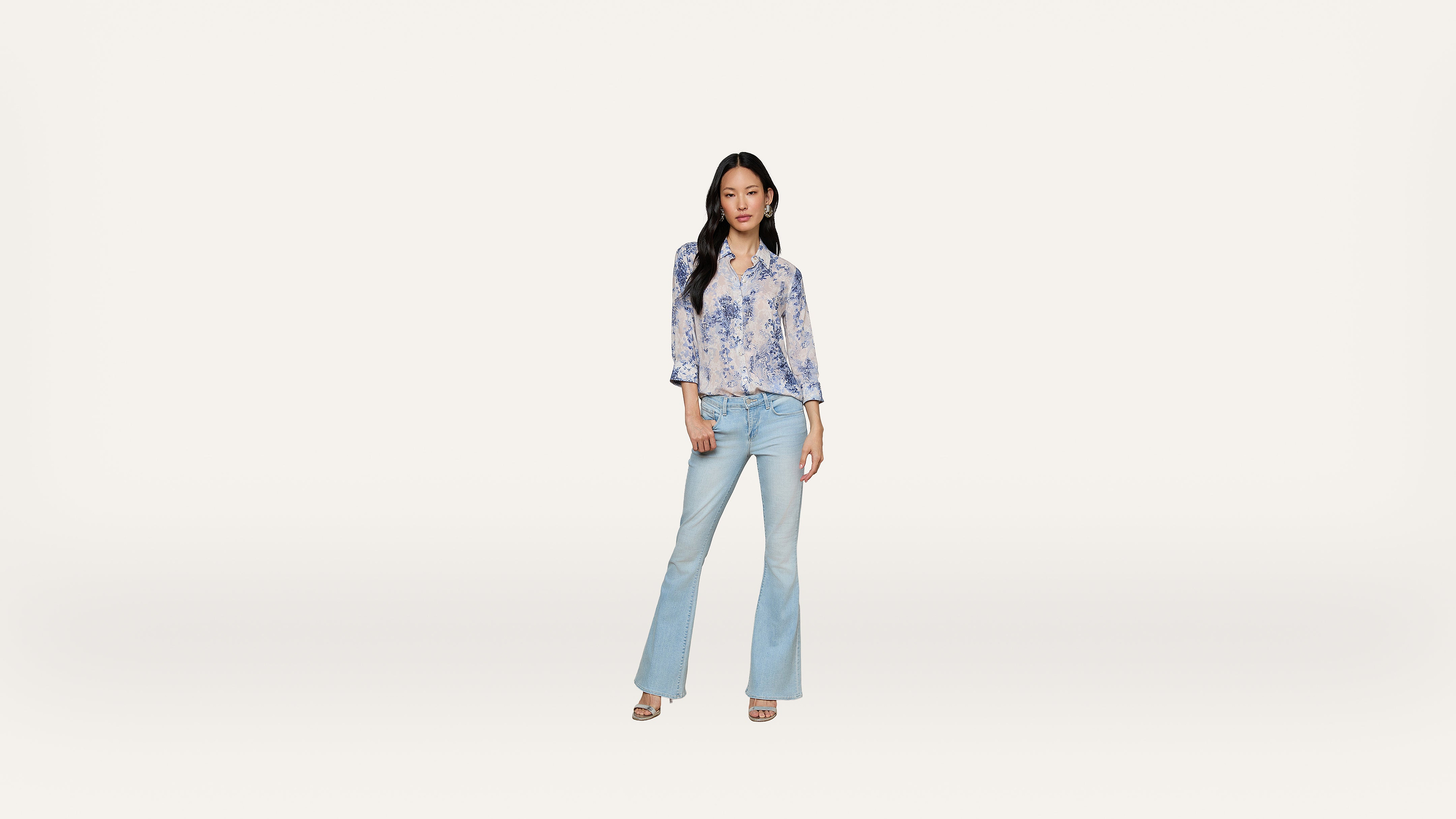 A woman stands against a plain white background, wearing a light blue floral blouse, light blue flared jeans, and sandals.