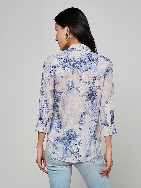 A woman with long black hair is seen from the back, wearing the Camille Blouse—an ivory and blue lace print three-quarter sleeve shirt crafted from viscose twill—paired with light blue jeans, set against a plain light background.