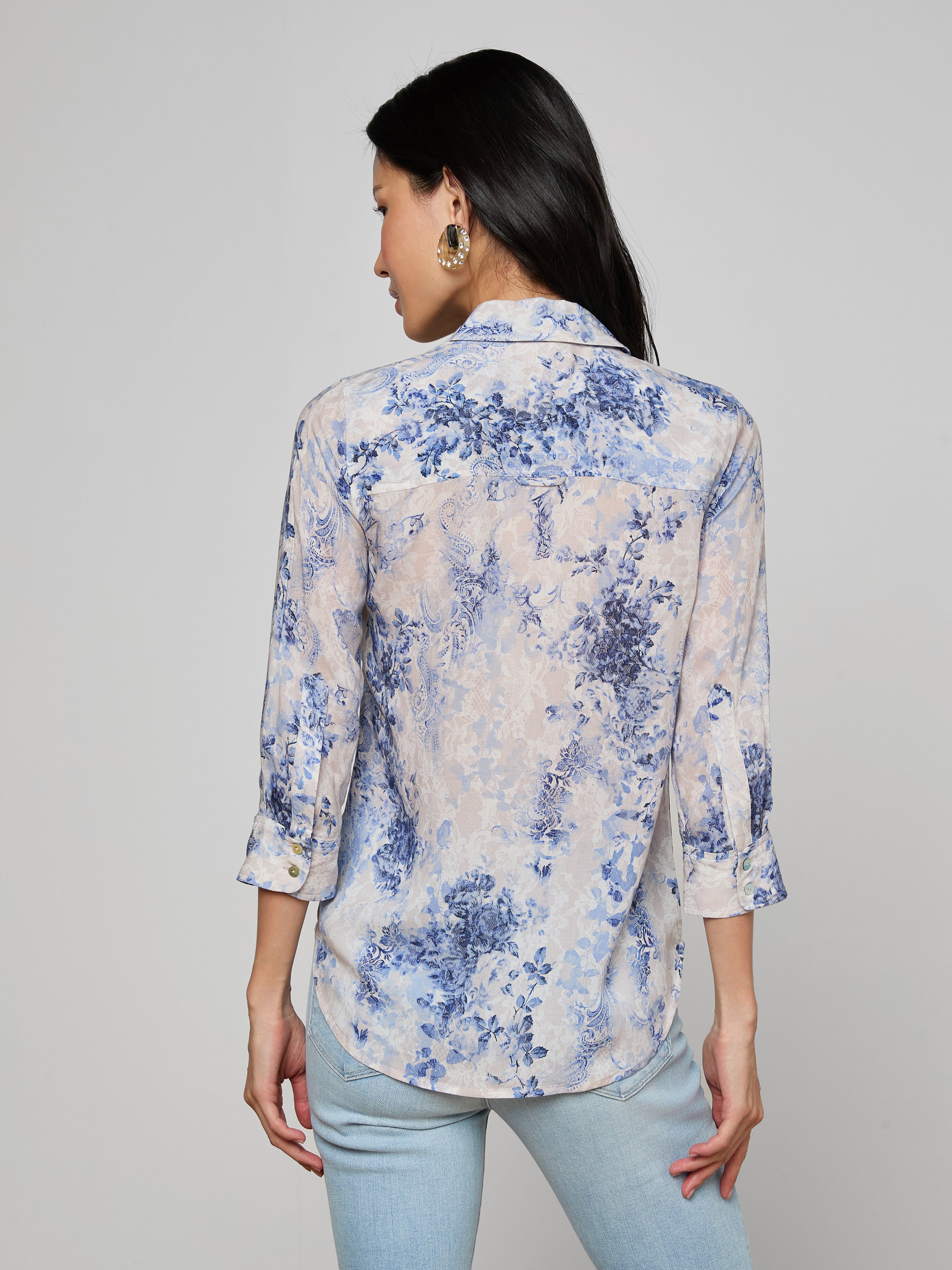 A woman with long black hair is seen from the back, wearing the Camille Blouse—an ivory and blue lace print three-quarter sleeve shirt crafted from viscose twill—paired with light blue jeans, set against a plain light background.