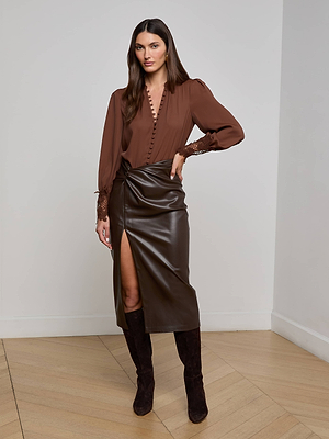 A woman stands indoors wearing the L'AGENCE Ava Blouse with scalloped lace cuffs, paired with a hot fudge brown faux leather wrap skirt featuring a front slit and knee-high brown suede boots. She poses with one hand on her waist.