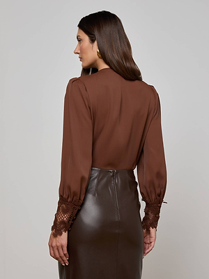 A woman faces away, wearing the L'AGENCE Ava Blouse in hot fudge brown with scalloped lace cuffs and a dark brown leather skirt. She has loose straight brown hair and gold hoop earrings against a plain light background.