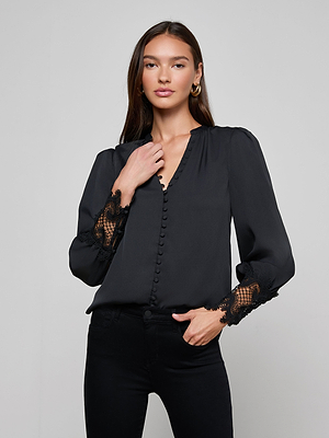 A woman with long brown hair models the L'AGENCE Ava Blouse, a vintage-inspired black top with scalloped lace sleeves, paired with black pants. She poses confidently against a plain background, one hand at her collar and the other in her pocket.