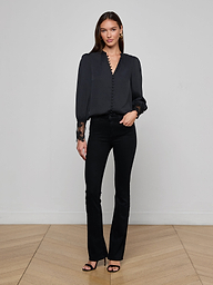 A woman stands against a plain white background wearing the Ava Blouse, styled with blue high-waisted jeans, a black belt featuring a gold buckle, and black heels.
