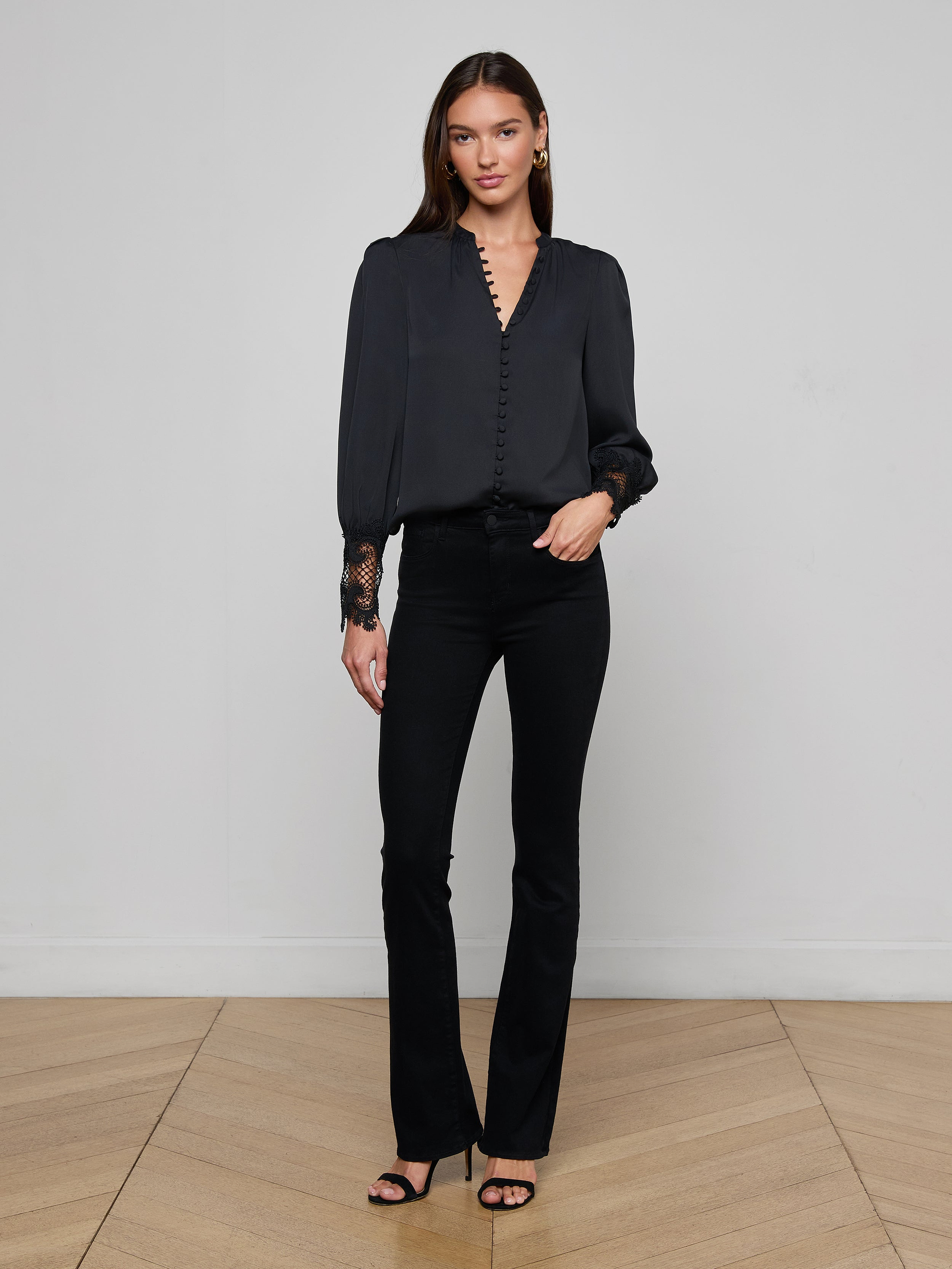 A woman stands against a plain white background wearing the Ava Blouse, styled with blue high-waisted jeans, a black belt featuring a gold buckle, and black heels.