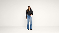 A woman stands against a plain white background, wearing a black long-sleeve top, blue wide-leg jeans, black heels, and a belt. She has long hair and is holding a phone in one hand.